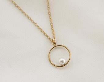 May include: A gold chain necklace with a delicate circle pendant featuring a single white pearl.