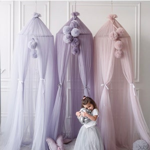 May include: Three sheer canopy curtains in shades of lavender and pink, with pom-pom accents hanging from the top. A young girl is standing in front of the curtains, holding a silver balloon.