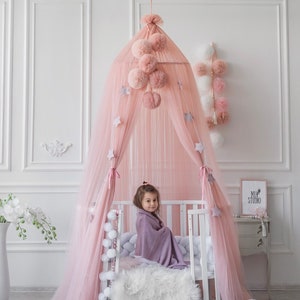 May include: A pink tulle canopy with white pom poms and stars hanging from it. A young girl is sitting in a white crib with a purple blanket. The crib is decorated with white pom poms and a white furry rug.