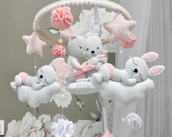 May include: A white and pink baby mobile with a wooden ring, clouds, stars, and bunnies. The bunnies are holding a smaller bunny. The stars are glittery.