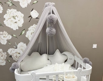 May include: White baby crib with a white canopy and gray pom-poms. The crib has a white mattress and a white blanket. There is a white bunny-shaped chair in front of the crib.