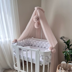 May include: White baby crib with a pink tulle canopy and a white braided bumper. The crib is on a white rug with a brown wooden floor. There is a wicker basket with stuffed animals next to the crib.
