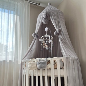 May include: A white crib with a gray canopy and a mobile hanging above it. The crib has a white mattress and a white bumper. The canopy is made of a sheer fabric and has a pom-pom trim. The mobile has a variety of toys hanging from it, including clouds, stars, and animals.
