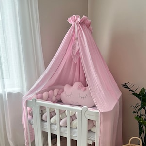 May include: A white cot with a pink canopy and a pink cloud-shaped pillow with a bow. The cot has a white bumper with pink, white, and grey braided rope.