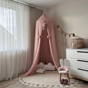 May include: A pink canopy with pom poms hangs from the ceiling over a white cloud-shaped pillow and a white chair with bunny ears. A round rug with fringe is on the floor.