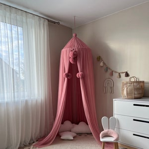 May include: A pink tulle canopy with pom-poms hangs over a round rug with tassels. There are white pillows and a white bunny chair inside the canopy.