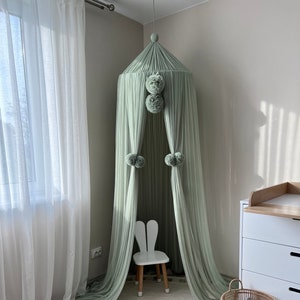 May include: A light green tulle canopy with pom poms hangs over a white bunny-shaped chair and a round jute rug with fringe. The canopy is in a room with white walls and a white dresser.