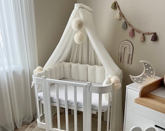 May include: White baby crib with a white canopy and a white bumper. The crib has a white mattress and a white sheet. The crib is in a room with a white wall and a white rug. There are some decorations on the wall, including a rainbow and a moon.