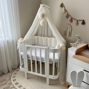 Könnte beinhalten: Ein weißes Babybett mit einem weißen Baldachin und einem weißen Stoßfänger. Das Bett hat eine weiße Matratze und ein weißes Laken. Das Bett steht in einem Raum mit einer weißen Wand und einem weißen Teppich. An der Wand befinden sich einige Dekorationen, darunter ein Regenbogen und ein Mond.