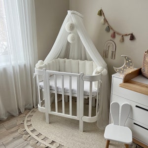 May include: White baby cot with a white canopy and a white bunny chair. The cot has a white mattress and a white bumper. The room is decorated with a white rug and a moon-shaped nightlight.