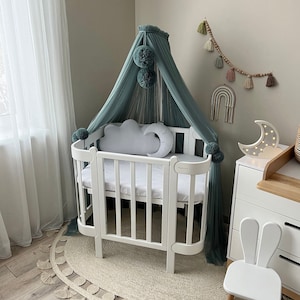 Puede incluir: Cuna blanca para bebé con una almohada en forma de nube blanca y un dosel turquesa con pompones. La cuna está sobre una alfombra beige con un borde de flecos. Hay una luz nocturna blanca con forma de luna creciente en una cómoda blanca.