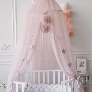 May include: A pink tulle canopy with a white pom-pom trim hangs over a white crib with a white braided bumper. The canopy is decorated with silver stars and pink pom-poms.