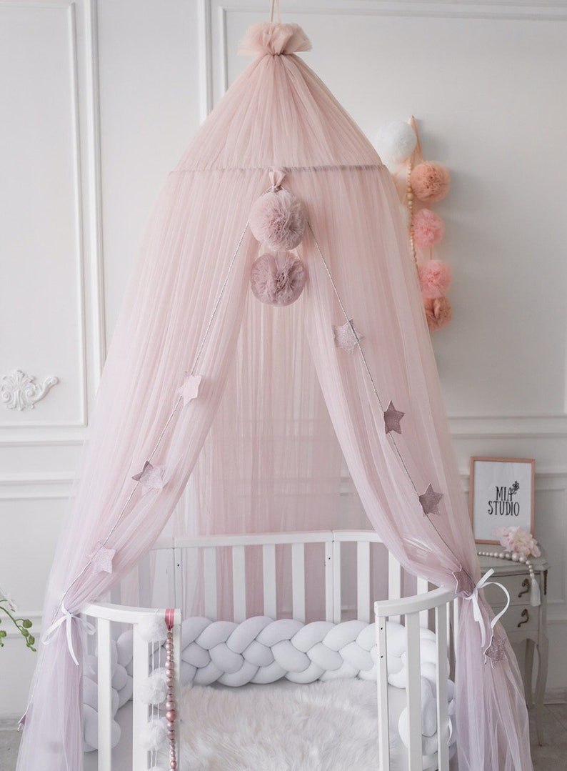 May include: A pink tulle canopy with a white pom-pom trim hangs over a white crib with a white braided bumper. The canopy is decorated with silver stars and pink pom-poms.