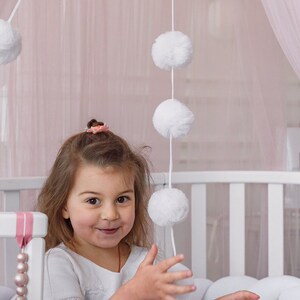 May include: A young girl wearing a white shirt with lace trim and a gray tulle skirt is playing with a white pom-pom garland hanging from a pink canopy. The girl is smiling and looking at the camera.