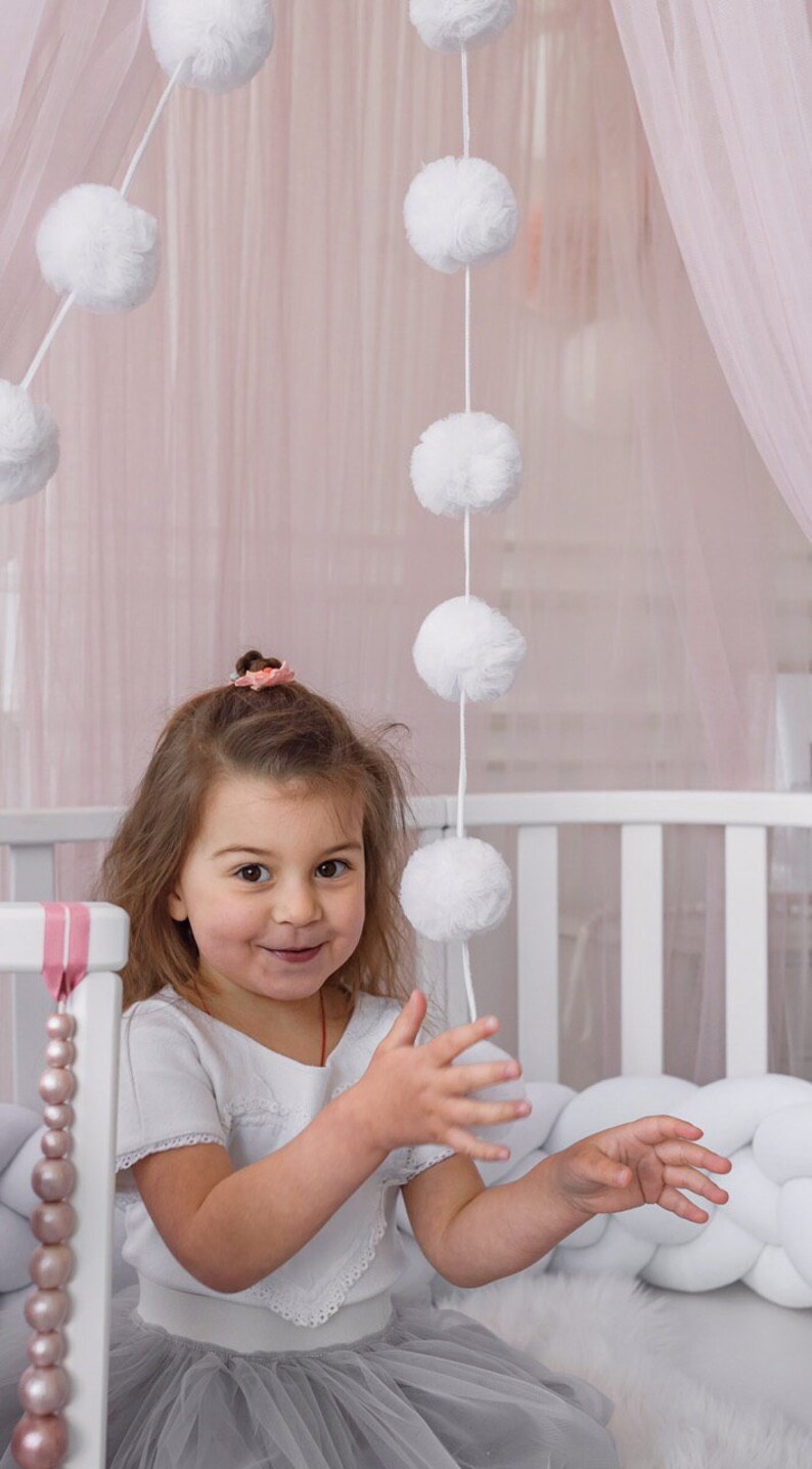 May include: A young girl wearing a white shirt with lace trim and a gray tulle skirt is playing with a white pom-pom garland hanging from a pink canopy. The girl is smiling and looking at the camera.