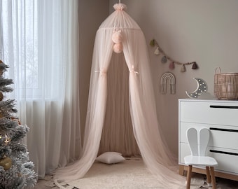 Könnte beinhalten: Ein hellrosa Tüllbaldachin mit weißen Pompons hängt von der Decke. Der Baldachin ist über einen runden Teppich mit Fransen drapiert. Ein weißer Stuhl mit Hasenohren steht vor dem Baldachin.
