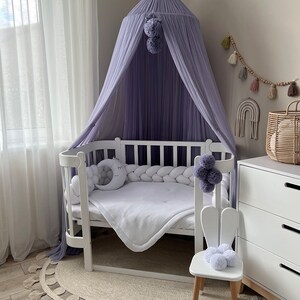 May include: A white crib with a purple canopy and a white bunny chair. The crib has a white mattress and a white blanket. The canopy is made of a sheer fabric and has a pom-pom trim. The bunny chair has a white seat and wooden legs.