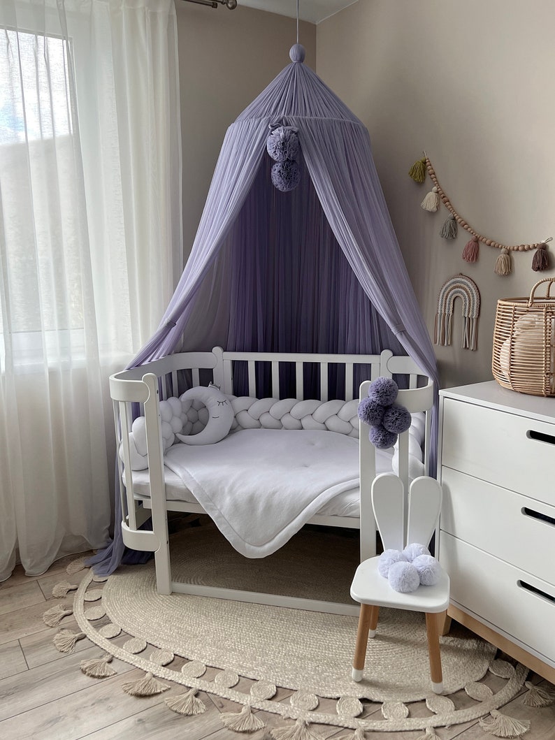 May include: A white crib with a purple canopy and a white bunny chair. The crib has a white mattress and a white blanket. The canopy is made of a sheer fabric and has a pom-pom trim. The bunny chair has a white seat and wooden legs.