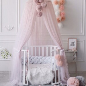 May include: A pink tulle canopy with pom-poms hangs over a white crib with a white braided bumper. A gray round rug with a white ruffle is on the floor. There are several pink pom-poms on the floor and a white furry rug in the crib.