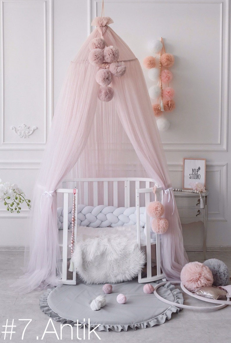 May include: A pink tulle canopy with pom-poms hangs over a white crib with a white braided bumper. A gray round rug with a white ruffle is on the floor. There are several pink pom-poms on the floor and a white furry rug in the crib.