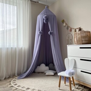 May include: A lavender-colored canopy with pom-poms hangs from the ceiling over a round jute rug with fringe. A white bunny-shaped chair with a gray pom-pom sits on the rug.