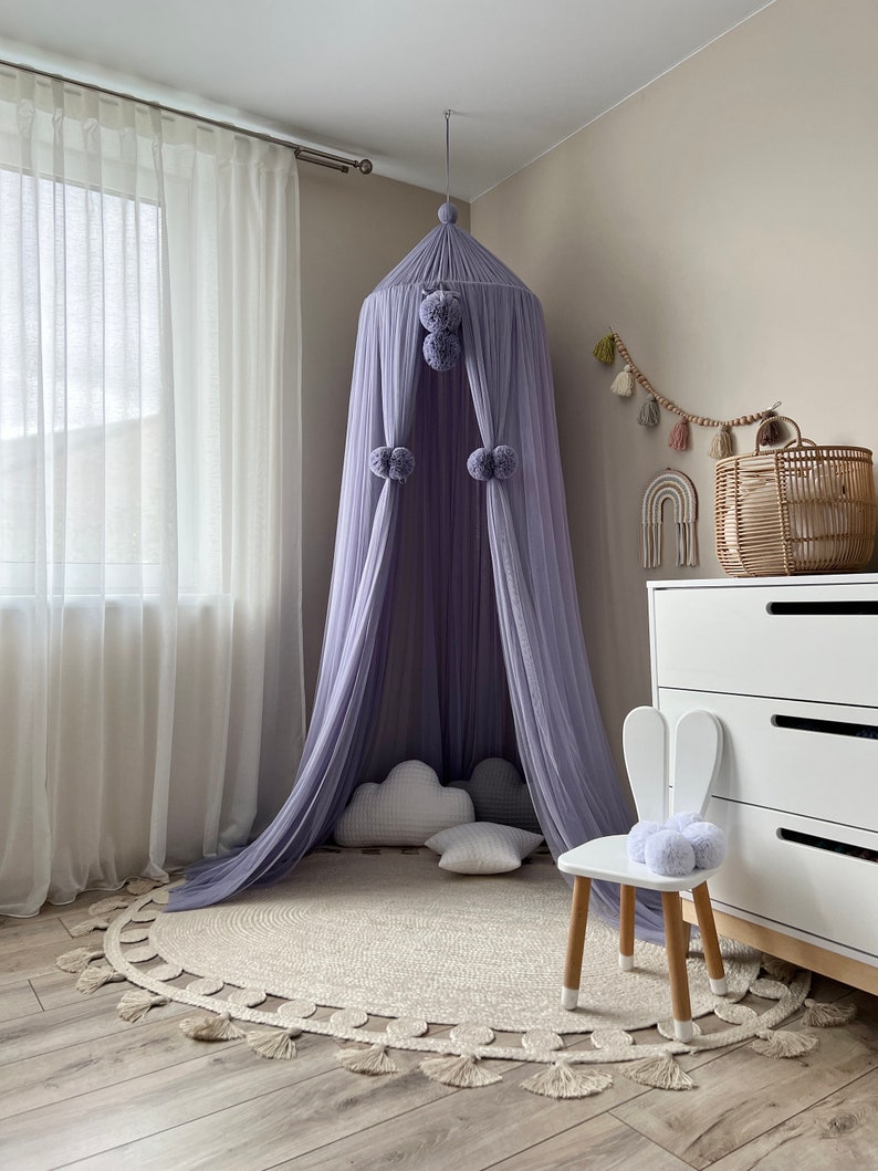 May include: A lavender-colored canopy with pom-poms hangs from the ceiling over a round jute rug with fringe. A white bunny-shaped chair with a gray pom-pom sits on the rug.