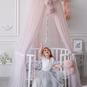 May include: A pink tulle canopy with pom-poms hangs over a white crib with a gray and white braided bumper. A young girl in a white shirt and a gray tulle skirt sits in the crib, reaching up towards the canopy. The crib is in a room with white walls and a white dresser with a sign that says "Mia Studio".