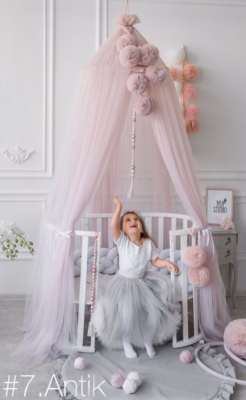 May include: A pink tulle canopy with pom-poms hangs over a white crib with a gray and white braided bumper. A young girl in a white shirt and a gray tulle skirt sits in the crib, reaching up towards the canopy. The crib is in a room with white walls and a white dresser with a sign that says "Mia Studio".
