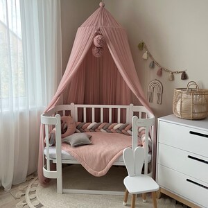 May include: A white crib with a pink canopy and a white bunny chair. The crib has a pink and white braided bumper and a pink blanket. The room is decorated with a white rug with tassels and a white dresser.