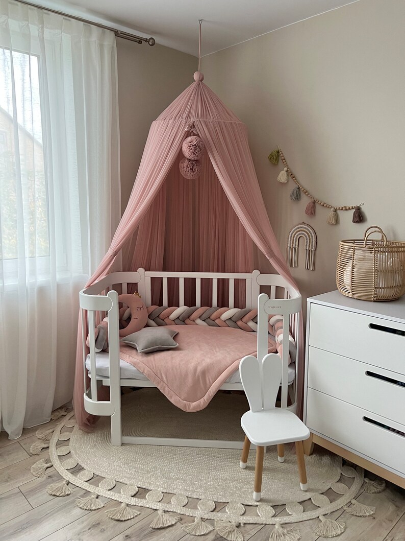 May include: A white crib with a pink canopy and a white bunny chair. The crib has a pink and white braided bumper and a pink blanket. The room is decorated with a white rug with tassels and a white dresser.