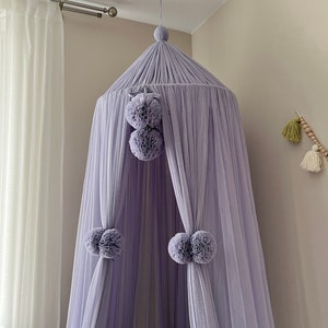 May include: A lavender-colored canopy bed with pom-pom accents. The canopy is made of a sheer fabric and hangs from the ceiling.