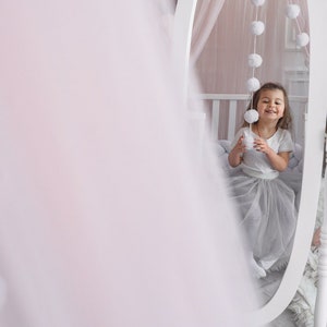May include: A young girl in a white shirt and a tulle skirt sits in front of a white oval mirror. The mirror is decorated with a string of white pom-poms. The girl is smiling and holding a white pom-pom.