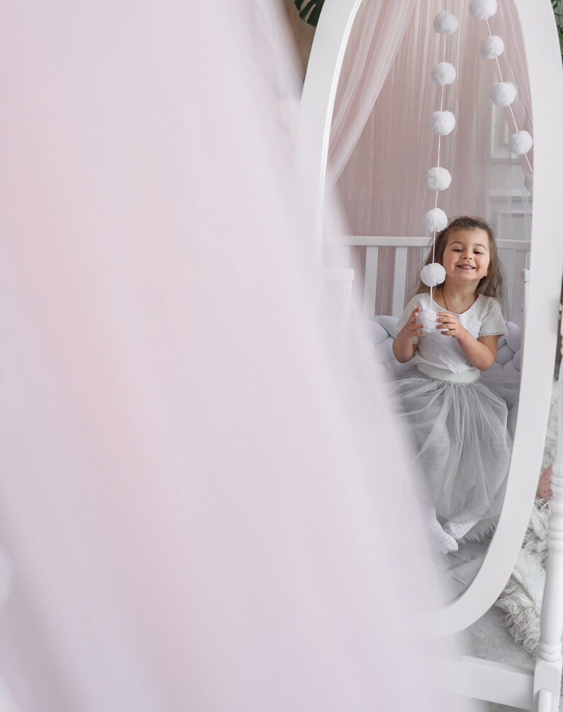May include: A young girl in a white shirt and a tulle skirt sits in front of a white oval mirror. The mirror is decorated with a string of white pom-poms. The girl is smiling and holding a white pom-pom.