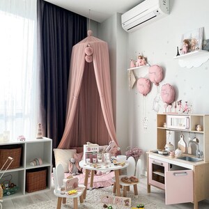 May include: A pink and white play kitchen with a pink oven and a sink. There are two white chairs with pink accents and a pink and white play food set on a white table. The kitchen is in a room with a pink canopy bed and a white rug.