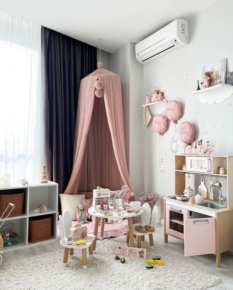 May include: A pink and white play kitchen with a pink oven and a sink. There are two white chairs with pink accents and a pink and white play food set on a white table. The kitchen is in a room with a pink canopy bed and a white rug.