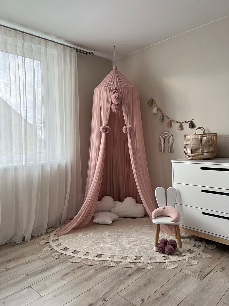May include: A pink canopy with pom poms hangs from the ceiling over a white cloud-shaped pillow and a white chair with bunny ears. A round rug with fringe is on the floor.