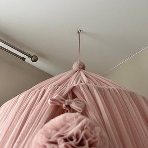 May include: A pink tulle canopy with a large pink pom-pom hanging from the center. The canopy is suspended from the ceiling with a white cord.