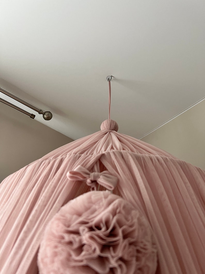 May include: A pink tulle canopy with a large pink pom-pom hanging from the center. The canopy is suspended from the ceiling with a white cord.