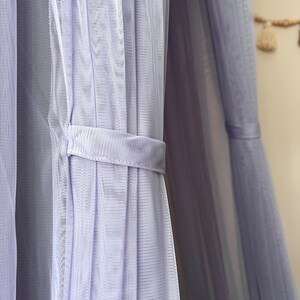 May include: A close-up of a lavender-colored sheer fabric curtain with a white ribbon tie. The fabric is gathered and pleated, creating a soft and delicate look.
