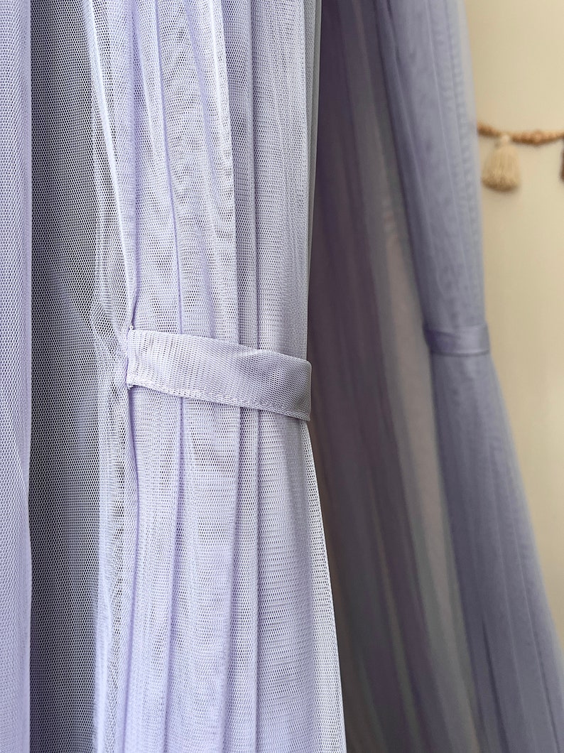 May include: A close-up of a lavender-colored sheer fabric curtain with a white ribbon tie. The fabric is gathered and pleated, creating a soft and delicate look.