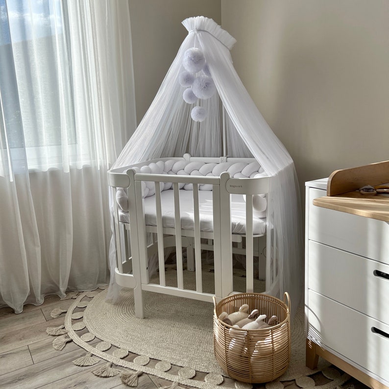 Puede incluir: Cuna blanca con dosel blanco y un m&oacute;vil de pompones blancos colgando encima. La cuna tiene un parachoques blanco y un colch&oacute;n blanco. Hay una c&oacute;moda blanca en el fondo y una cesta de mimbre con animales de peluche en primer plano.