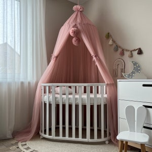 May include: A white oval crib with a pink tulle canopy and white pom poms. The crib is in a nursery with a white dresser and a white bunny rabbit chair.