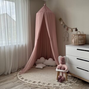 May include: A pink canopy bed with white pillows and a white rug with fringe. The bed is in a room with white curtains and a white dresser.