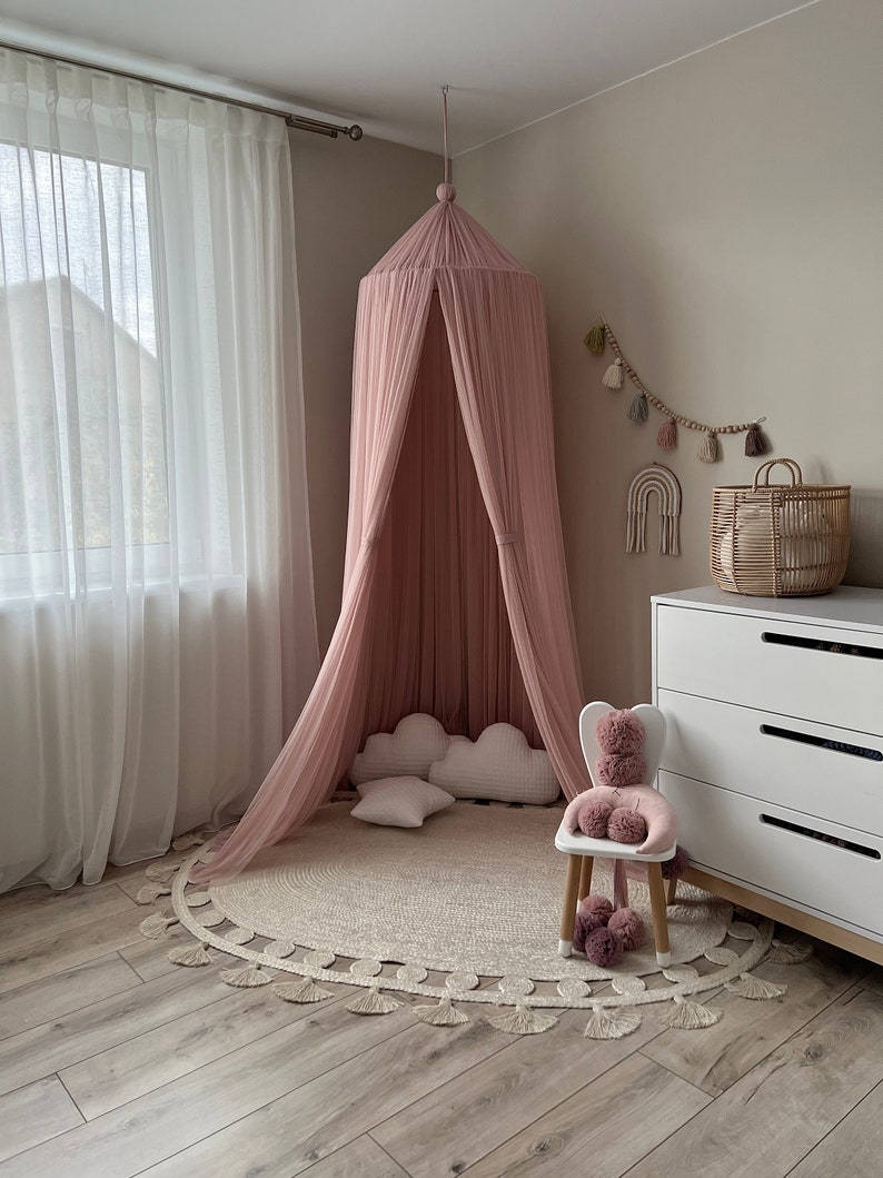 May include: A pink canopy bed with white pillows and a white rug with fringe. The bed is in a room with white curtains and a white dresser.
