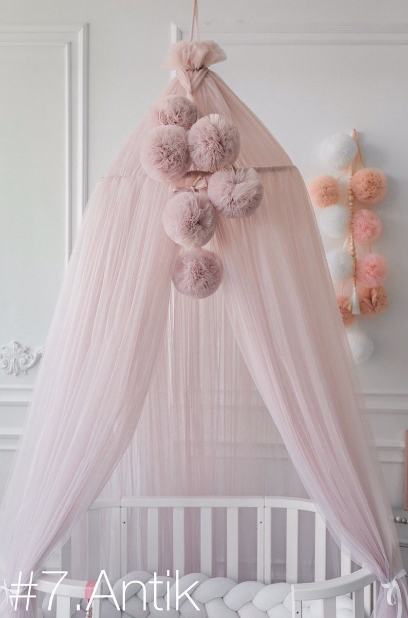 May include: A pink tulle canopy with a white crib underneath. The canopy has a cluster of pink pom-poms hanging from it. The text #7. Antik is visible in the bottom left corner of the image.