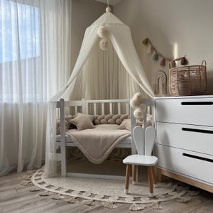 May include: A white crib with a white canopy and a beige blanket. A white bunny-shaped chair sits next to the crib. The crib is decorated with a braided rope bumper and a white pom-pom garland.