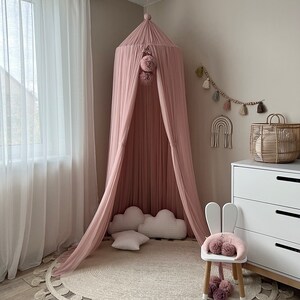 May include: A pink canopy with pom poms hangs from the ceiling above a white rug with a fringe border. A white bunny chair with pink pom poms sits on the rug.