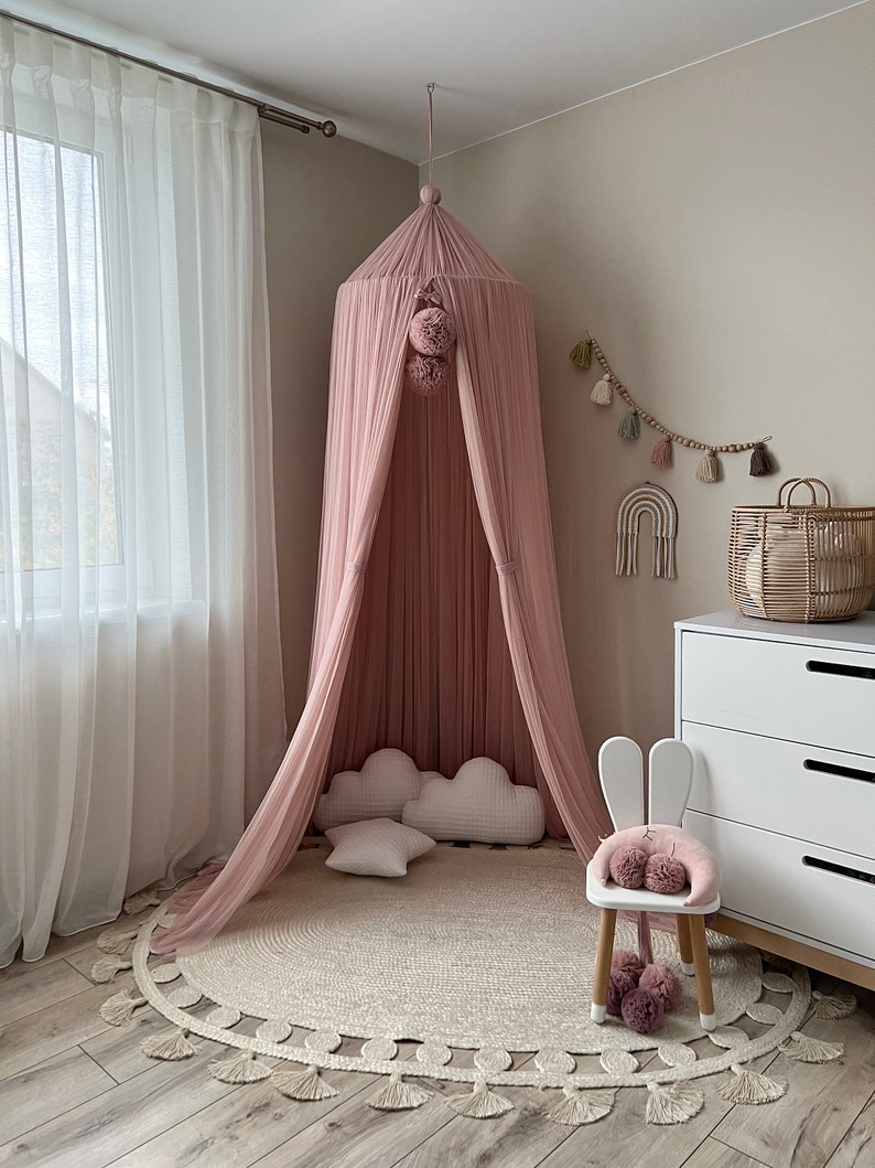 May include: A pink canopy with pom poms hangs from the ceiling above a white rug with a fringe border. A white bunny chair with pink pom poms sits on the rug.