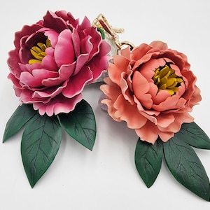 May include: Two handcrafted peony flower keychains. One is a deep pink, the other is coral, both with yellow centres and green leaves. The keychains have gold-coloured hardware and are set against a white background.