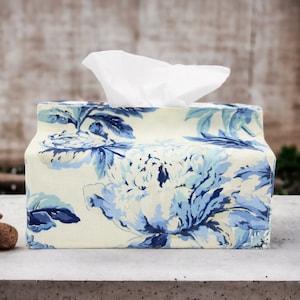 Op de afbeelding: Een witte tissueboxhoes met een blauw en wit bloemenpatroon. De hoes is gemaakt van stof en heeft een rechthoekige vorm.